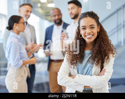 Ich bin immer bestrebt, Erfolg zu haben. Porträt einer jungen Geschäftsfrau, die in einem Büro mit ihren Kollegen im Hintergrund steht. Stockfoto