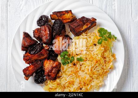 Nahaufnahme von geschmortem Sauerkraut mit Schweinefleisch und Pflaumen auf weißem Teller auf Holztisch, horizontale Ansicht von oben, flaches Lay Stockfoto