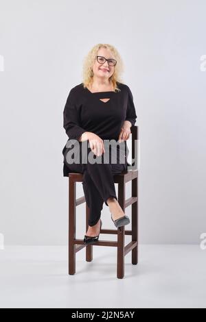 Stilvolle elegante Frau mittleren Alters mit blonden Haaren trägt schwarze Bluse und Hose sitzt auf Stuhl Stockfoto