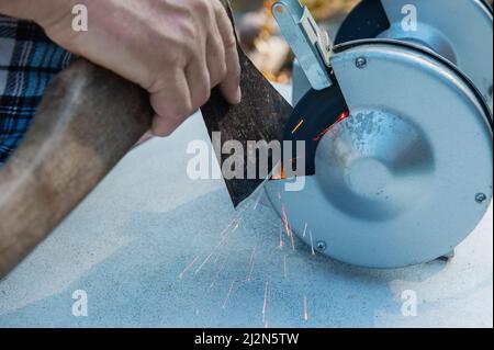 Manuelles Schärfen einer Axt mit Handwerkzeugen. Beschnittene Hände einer unbekannten Person Schärfen einer Axt auf Einer Schreibmaschine. Der Mensch schärft ein Werkzeug Stockfoto