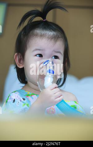 Nettes asiatisches Kind hält einen Maskendampf-Inhalator für die Behandlung von Asthma auf Krankenbett im Krankenhaus. Atmen durch einen Dampfvernebler. Konzept der Inhalation Stockfoto