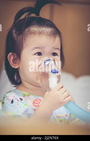 Nettes asiatisches Kind hält einen Maskendampf-Inhalator für die Behandlung von Asthma auf Krankenbett im Krankenhaus. Atmen durch einen Dampfvernebler. Konzept der Inhalation Stockfoto