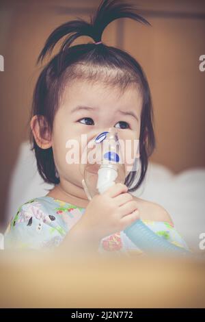 Nettes asiatisches Kind hält einen Maskendampf-Inhalator für die Behandlung von Asthma auf Krankenbett im Krankenhaus. Atmen durch einen Dampfvernebler. Konzept der Inhalation Stockfoto