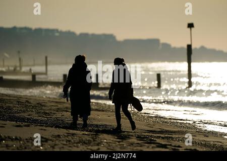 Die Menschen laufen am Strand entlang, während die Sonne über dem Strand von Boscombe in Dorset aufgeht. Bilddatum: Sonntag, 3. April 2022.
