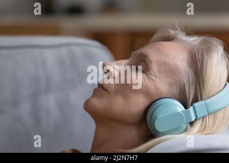 Friedliche, reife Dame in großen kabellosen Kopfhörern, die sich auf der Couch ausruhen Stockfoto