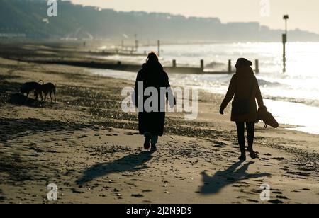 Die Menschen laufen am Strand entlang, während die Sonne über dem Strand von Boscombe in Dorset aufgeht. Bilddatum: Sonntag, 3. April 2022.