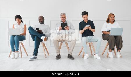 Ein paar lohnende Verbindungen herzustellen, während sie warten. Aufnahme einer vielfältigen Gruppe von Menschen, die digitale Geräte benutzen, während sie in einer Schlange gegen einen Weißen sitzen Stockfoto