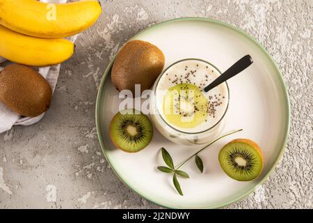 Hausgemachter, frisch zubereiteter Kiwi- und Bananenmoothie mit Chiasamen im Glas auf einem Keramikblech und betontem Hintergrund. Draufsicht. Reinigung des Körpers Stockfoto