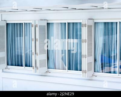 Weiße, klappbare Holzfenster mit Glasfenstern und blauen Vorhängen auf weißem Gebäude. Fenster im asiatischen Stil, Außendesign. Stockfoto