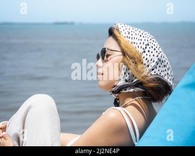 Schöne asiatische Frau Porträt mit geschlossenen Augen in lässigen weißen Hemd tragen Sonnenbrille und Haarschal sitzen auf blauen Sitzsack am Strand mit s Stockfoto