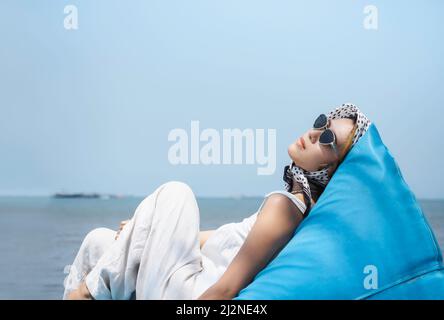 Schöne asiatische Frau in lässigem weißen Hemd und Hose trägt Sonnenbrille und Haarschal auf blauen Sitzsack mit Blick auf das Meer und blauen Himmel liegend Stockfoto
