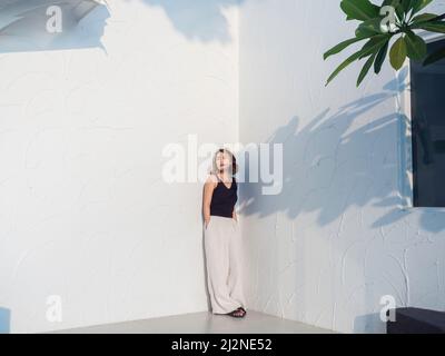 Asiatische Frau sieht cool aus, mit kurzen Haaren, die ein schwarzes Tanktop und eine weiße Hose tragen und die Hände in den Taschen auf weißem Hintergrund halten, voll Stockfoto