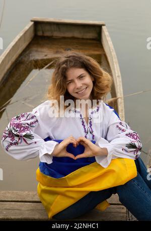 Schönes Mädchen 17-19 Jahre alt in besticktem Hemd mit ukrainischer Flagge am Flussufer. Die Hände sind in Form des Herzens gefaltet. Die Ukrainer sind gegen den Krieg. Stockfoto