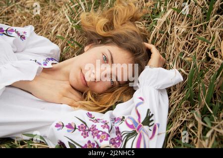 Schönes Mädchen 17-19 Jahre alt in einem gestickten ukrainischen traditionellen Hemd liegt in trockenem Gras. Blick von oben. Ich bin Ukrainerin. Stehe mit der Ukraine Stockfoto