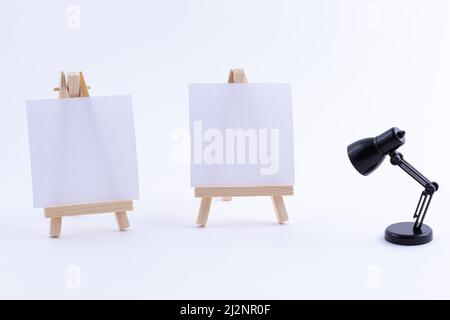 Zwei hölzerne Easels Miniature mit blanker weißer quadratischer Leinwand für Künstler und Maler - Mockup. Mini-Holzständer mit sauberer Zeichenfläche und kleiner schwarzer Tischleuchte auf weißem Hintergrund, Kopierraum Stockfoto