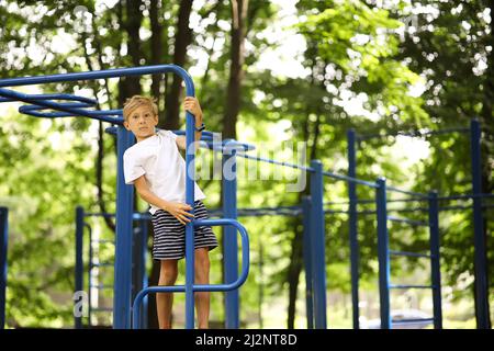 Der Athlet Boy im Park geht zum Sport, der an seinen Händen hängt und macht mit gekreuzten Beinen Übungen an den unebenen Riegel Stockfoto