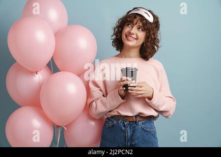 Glückliche Frau in Schlafmaske trinken Kaffee, während posiert mit rosa Ballons isoliert auf blauem Hintergrund Stockfoto