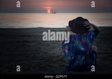 Frau in einem Hut sitzt am Strand und schaut auf den Sonnenaufgang am Meer Stockfoto