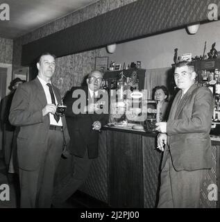 1963, historisch, drei von mir stehen, trinken und rauchen in einer Bar, Stockport, Manchester, England, Großbritannien. Glashülle an der Bar mit Schweinekuchen und Schild für die Jubliee-Biermarke, am bekanntesten ein Jubliee Stout. Jubliee Beer wurde von Hope and Anchor of Sheffield gebraut, die 1962 mit der Charington-Brauerei in Mile End, London, fusionierten. Stockfoto