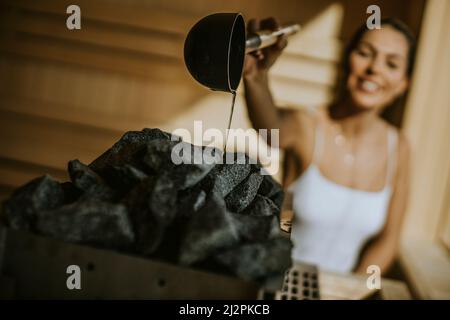 Attraktive junge Frau, die in der Sauna Wasser auf heißen Stein gießt Stockfoto