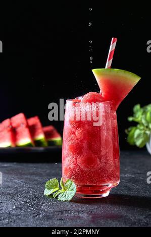 Reife Wassermelonen-Noisette-Kugeln mit sada im Glas und fallenden Wassertropfen auf dunklem Hintergrund. Erfrischender Sommerdrink Stockfoto