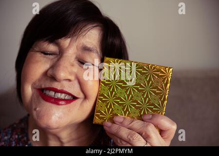 Eine ältere Frau, die mit geschlossenen Augen lächelt und ein gelbes Geschenk in der Schachtel hält Stockfoto