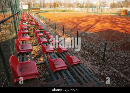 Details des Tennisplatzes der Stadt im Winter, rote leere Stühle und viele heruntergefallene Blätter an einem sonnigen Tag auf dem Platz. Stockfoto