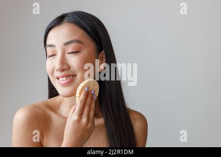 Fröhliche asiatische Frau Reinigung Gesicht mit kosmetischen Schwamm Stockfoto