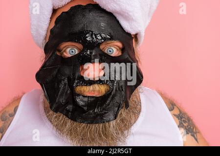 Zweifler Mann mit Bart verhält sich wie eine Frau und hat Spaß mit einer Gesichtsmaske Stockfoto
