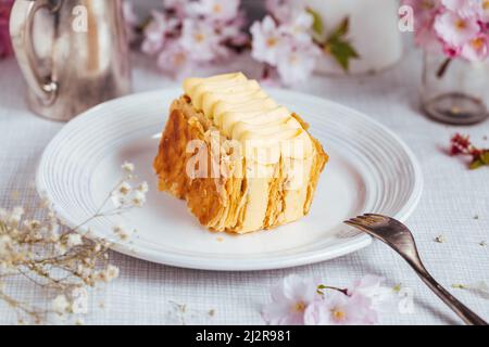 Französischer Nachtisch namens Mille Feuille oder Napoleon-Kuchen, verziert mit Blütenblumen auf hellem Hintergrund. Geschichteter leckerer Kuchen mit Vanillecreme und pu Stockfoto