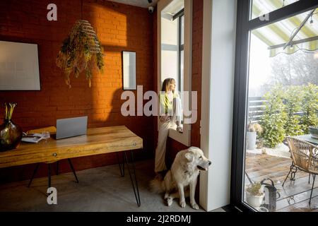 Frau, die auf der Fensterbank sitzt und mit ihrem weißen, entzückenden Hund zu Hause aus dem Fenster schaut Stockfoto