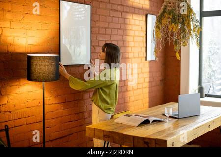 Eine Frau hängt zu Hause einen Fotorahmen an einer Ziegelwand Stockfoto
