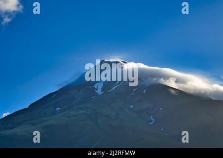 Vulkan mit Schneefeldern und Wolken rund um den Gipfel, beleuchtet von der Abendsonne. Mount Taranaki (Mount Egmont), Nordinsel, Neuseeland. Stockfoto