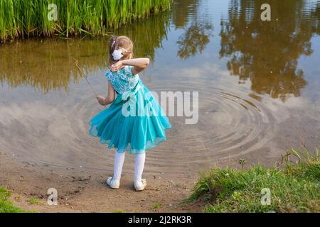 Ein Mädchen in einem blauen Kleid wirft Kieselsteine ins Wasser. Ein Kind spielt mit dem Fluss in der Natur. Ein elegant gekleidetes Baby hat Spaß in der Nähe des Sees. Chil Stockfoto