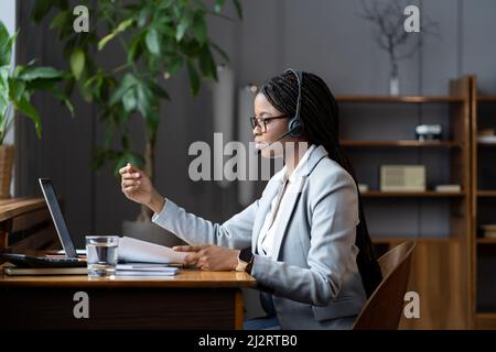 Selbstbewusste Geschäftsfrau im Headset hält bei Online-Videobesprechung Dokumente über das Projekt Stockfoto