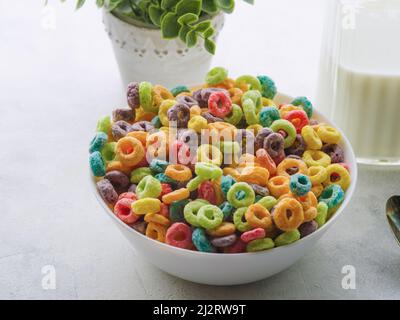 Schüssel mit mehrfarbigen Getreidefruchtringen, Milch in einem Krug und Dekor - Innenblume auf weißem Hintergrund. Nahaufnahme. Schnelles Vollkornfrühstück, expr Stockfoto