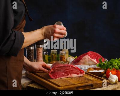 Der professionelle Küchenchef bereitet saftige Steaks aus frischem Fleisch auf einem Holzbrett mit dunklem Hintergrund zu. Die Fleischstücke mit Gewürzen bestreuen. Viele von mir Stockfoto