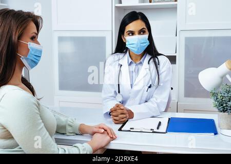 Arzt und Patient in Gesichtsmasken diskutieren Gesundheitsprobleme, während sie am Tisch in medizinischen Zentren im Gesundheitswesen sitzen Stockfoto