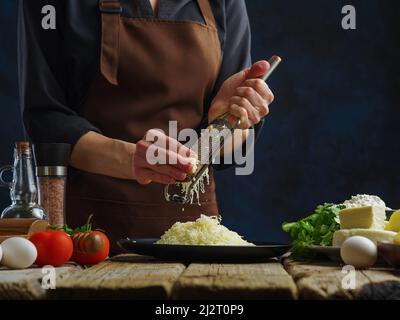 Der professionelle Koch reibt Käse auf einer manuellen Reibe auf einem Holztisch auf dunkelblauem Hintergrund. Gemüse, Kräuter, Gewürze stehen auf dem Tisch. Konzept - Rezept Stockfoto