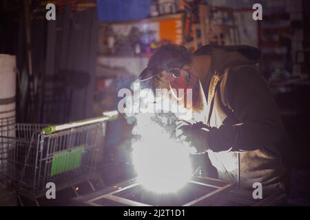 Metallschweißen. Arbeiten in der Werkstatt. Schweißer erzeugt Naht. Stahlverbindung. Mann in der Garage. Hohes Fieber. Schutz des Gesichts vor Flammen. Stockfoto