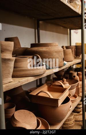 Regale mit Schüsseln, Tellern, Gläsern, Teekannen usw. aus Ton in natürlicher Rohfarbe bis zum endgültigen Gemälde. Kunsthandwerk aus Lehm. Vertikale Ausrichtung. Stockfoto