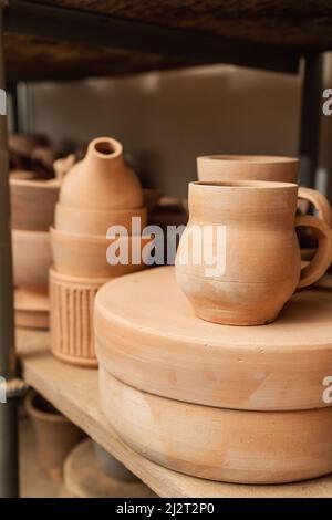 Regale mit Schüsseln, Tellern, Gläsern, Teekannen usw. aus Ton in natürlicher Rohfarbe bis zum endgültigen Gemälde. Kunsthandwerk aus Lehm. Vertikale Ausrichtung. Stockfoto