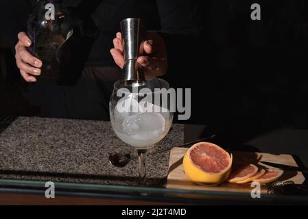 COCKTAIL WIRD ZUBEREITET. MESSUNG DER MENGE DES ALKOHOLISCHEN GETRÄNKS. GRAPEFRUITSCHEIBEN SCHNEIDEN. Stockfoto