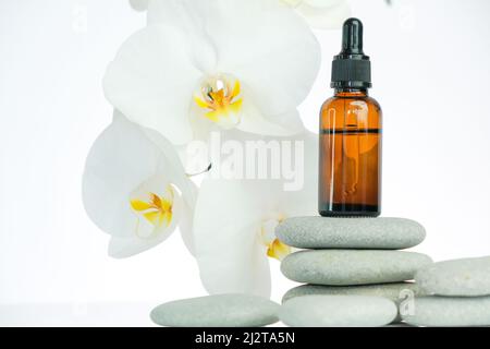 Massageöl und Massagesteine. Glasflasche mit Massageöl auf grauen Steinen und Orchideenblume auf weißem Hintergrund.Spa und Aromatherapie.Schönheit und Stockfoto