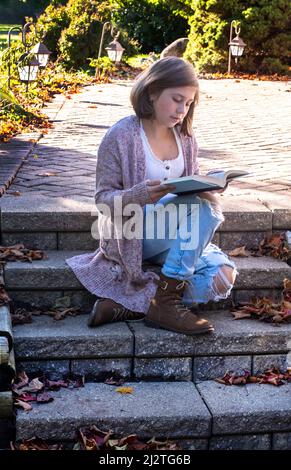 Ein junges Mädchen sitzt auf der Veranda und liest an einem schönen Herbsttag ein Buch Stockfoto