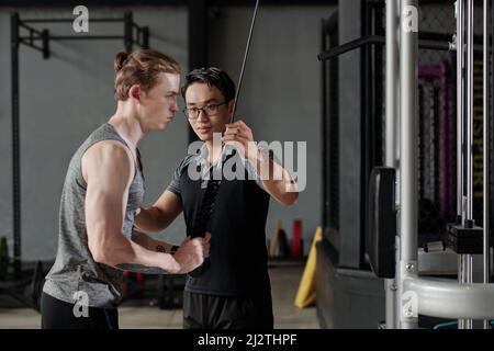 Fitness-Trainer Korrektur muskulösen jungen Mann dabei Kabel Pull-Down-Übung für Trizepmuskeln Stockfoto
