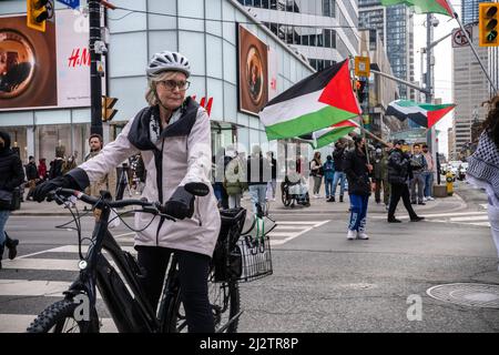 Toronto, Kanada. 02. April 2022. Eine Frau auf einem Fahrrad überquert die Straße, während Demonstranten mit palästinensischen Fahnen hinter ihr herlaufen. Demonstranten versammelten sich auf dem Yonge-Dundas-Platz in Toronto, Kanada, in Solidarität mit Palästina. Kredit: SOPA Images Limited/Alamy Live Nachrichten Stockfoto