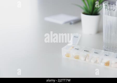 Nahaufnahme der medizinischen Pille Box mit Dosen von Tabletten für den täglichen Nehmen Sie ein Medikament mit weißen, gelben Drogen und Kapseln zur Behandlung verwendet, die Krankheit zu heilen. Glas Tasse Wasser auf dem Tisch, grauer Hintergrund. Stockfoto