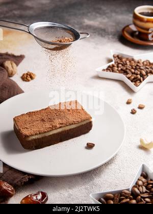 Schwarzer Kaffee in brauner kleiner Tasse, Kaffeebohnen in sternförmigen Tellern und gesunder zuckerfreier hausgemachter, roher veganer Tiramisu-Kuchen mit Cashewnüssen, Datteln in Stockfoto