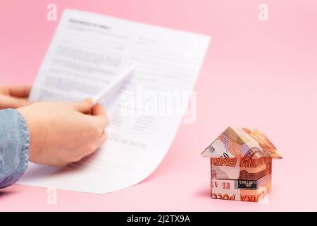 Ein kleines Origami-Haus aus russischen Rubel. Im Hintergrund zeigen die unscharfen Hände der Frauen den Vertrag. Rosa Hintergrund. Hypothekenkonzept. Stockfoto